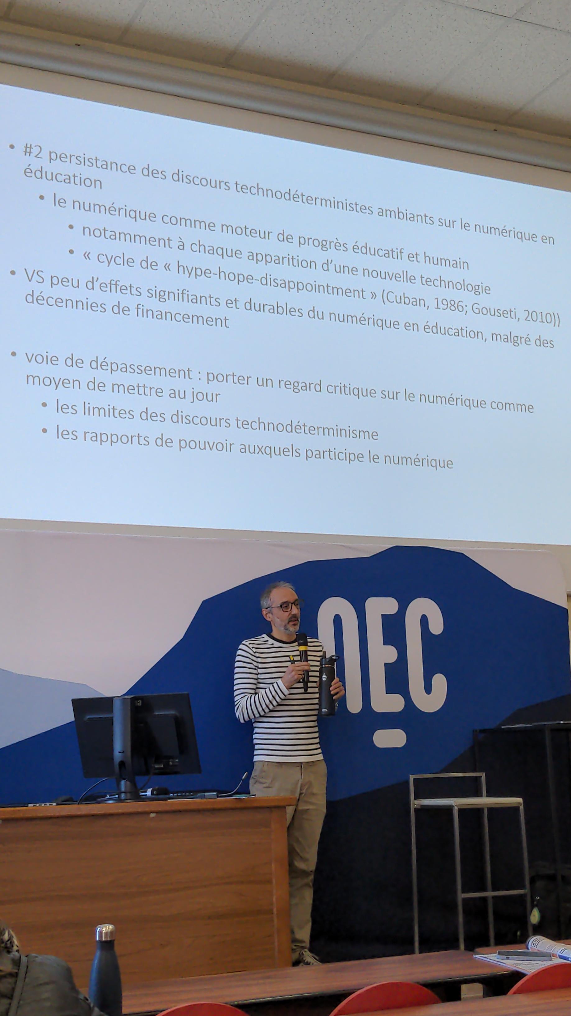 Le conférencier, Simon Collin, est debout sur l'estrade, devant un grand panneau NEC. Au-dessus de lui, sur le mur, est projetée une diapositive de son support de présentation.