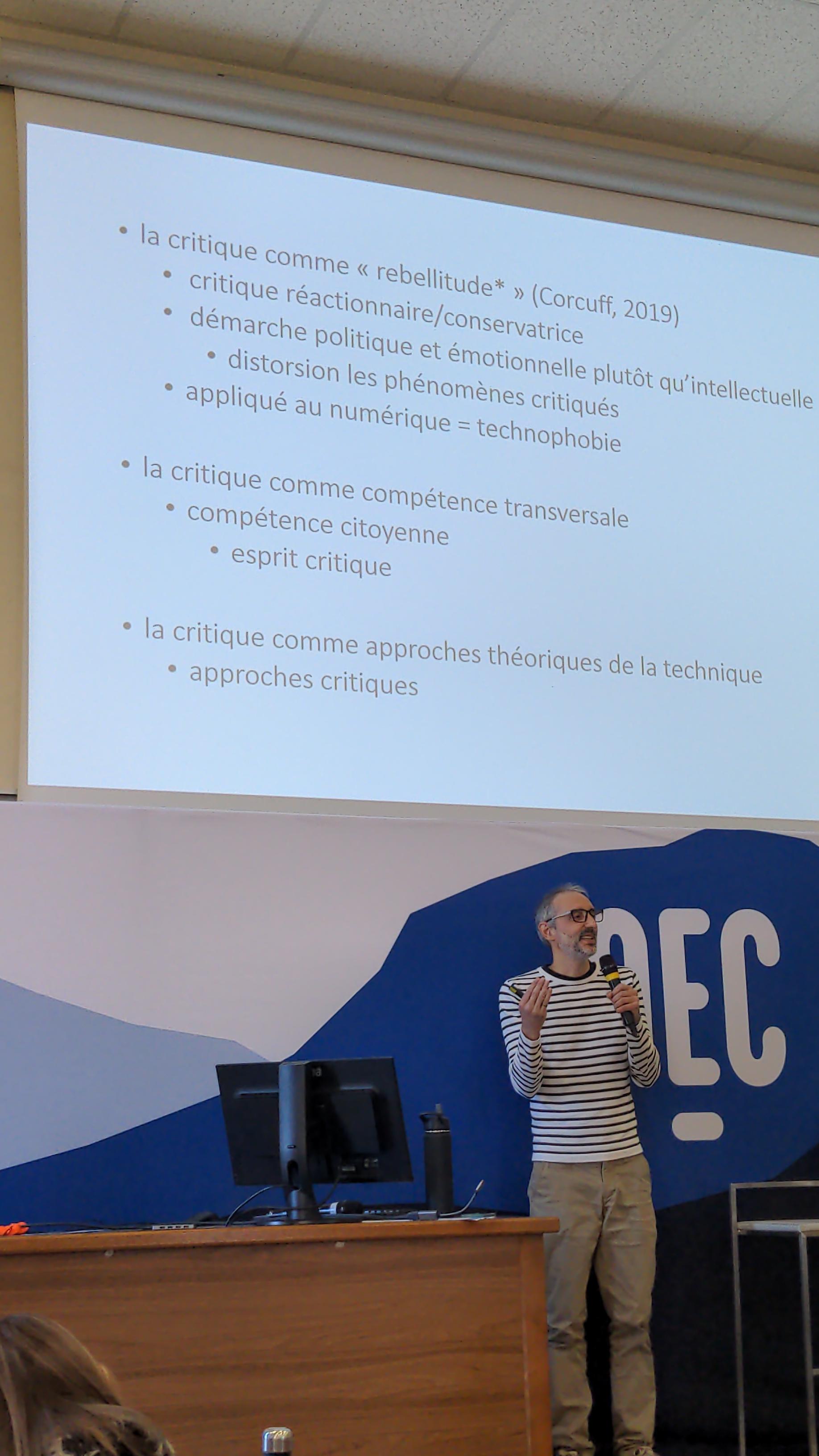 Le conférencier, Simon Collin, est debout sur l'estrade, et parle dans un micro, devant un grand panneau NEC. Au-dessus de lui, sur le mur, est projetée une diapositive de son support de présentation.