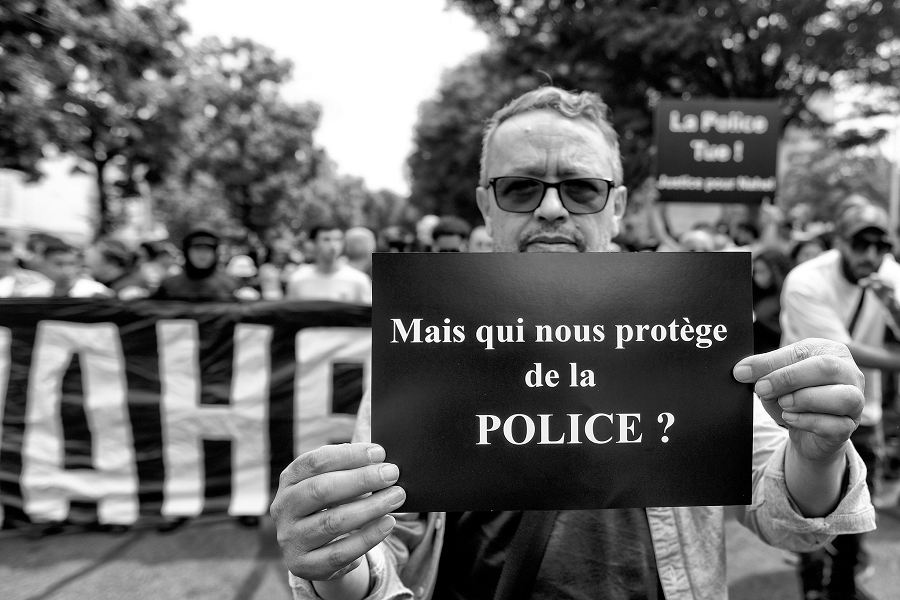 Un manifestant tient une feuille noire sur laquelle est inscrit "Mais qui nous protège de la police".