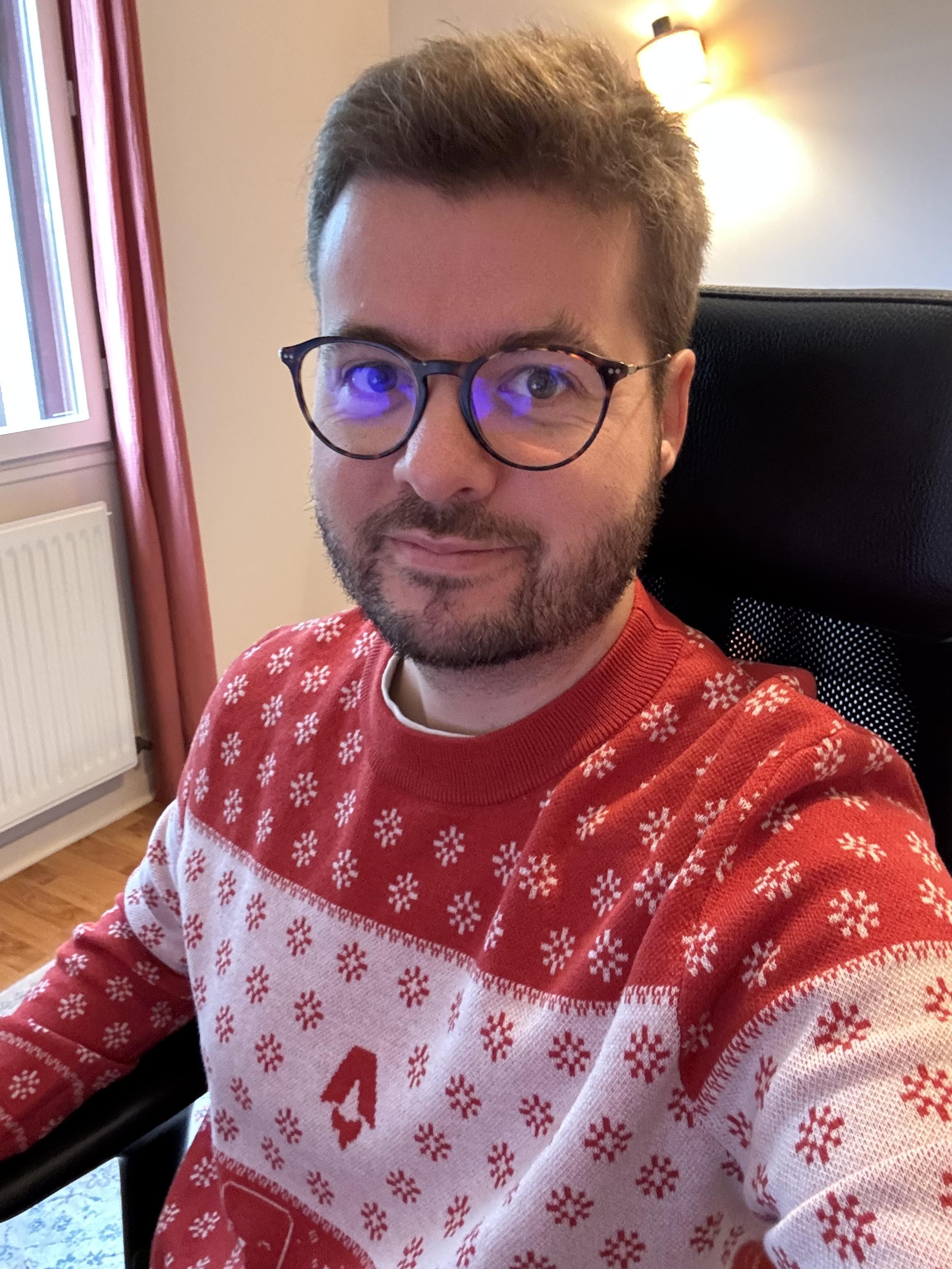 A picture of me in a Christmas jumper. The jumper is red and white. It sports the Astro logo on the chest. Starlight logos make up snowflakes. There are Houstons, and pixel art computers and planets.