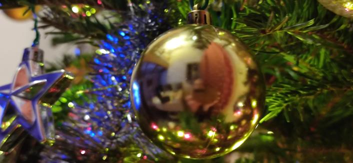 Gros plan sur une boule de Noël dorée dans un sapin décoré et éclairé. Scintillements multicolores. Une étoile argentée à gauche. Sur la surface de la boule dorée, on devine un visage derrière un smartphone en position paysage.