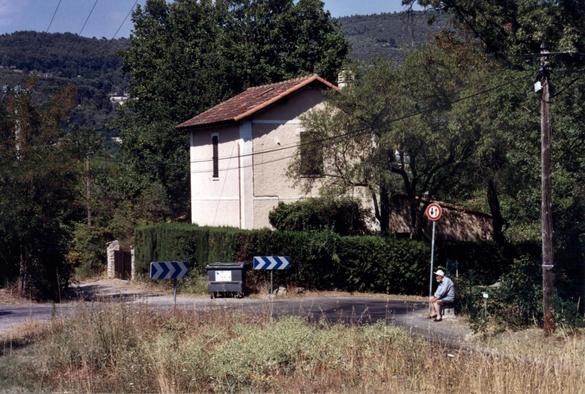 À distance, une maison aux volets clos et toiture de tuiles sous le ciel bleu. Elle est entourée d'une haie défensive. Devant, au milieu, un conteneur à poubelles entre deux panneaux de signalisation de virage. Un croisement de route. Un panneau indiquant que les véhicules en face ont la priorité.Une ligne électrique basse tension. Et sur la droite, au bord de la route, assis sur un muret, un homme âgé, le dos courbé, portant une casquette, une chemise bleue, un short et des espadrilles. Il est…
