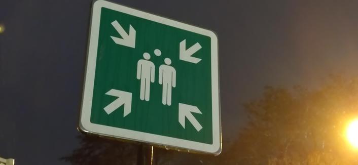 panneau vert et blanc indiquant un point de rassemblement. quatre flèches blanches partant des quatre coins du panneau carré pointent le centre où sont figurés trois silhouettes. c'est la nuit, un lampadaire éclaire l'image.