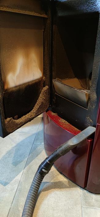 la porte du poêle à pellets rouge est grande ouverte, dévoilant une grande accumulation de cendres et de suie sur la paroi vitrée. un tuyau d'aspirateur équipé dun embout suceur s'apprête à tout faire disparaître.