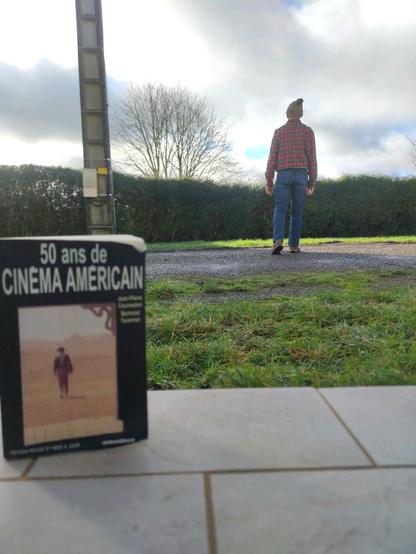 photo prise depuis une porte fenêtre. on voit à quelques mètres de là, un homme portant bonnet ridicule, chemise longue, ceinture et jeans, s'éloignant de la porte fenêtre dans un paysage verdoyant et humide pour la saison. Au premier plan, un bouquin se tient debout sur la margelle carrelée, on y voit une illustration d'un homme sorti d'un western américain, chapeau de cow-boy, chemise longue, bretelles et jeans, s'éloignant dans un paysage aride. au premier plan de l'illustration, on devine u…