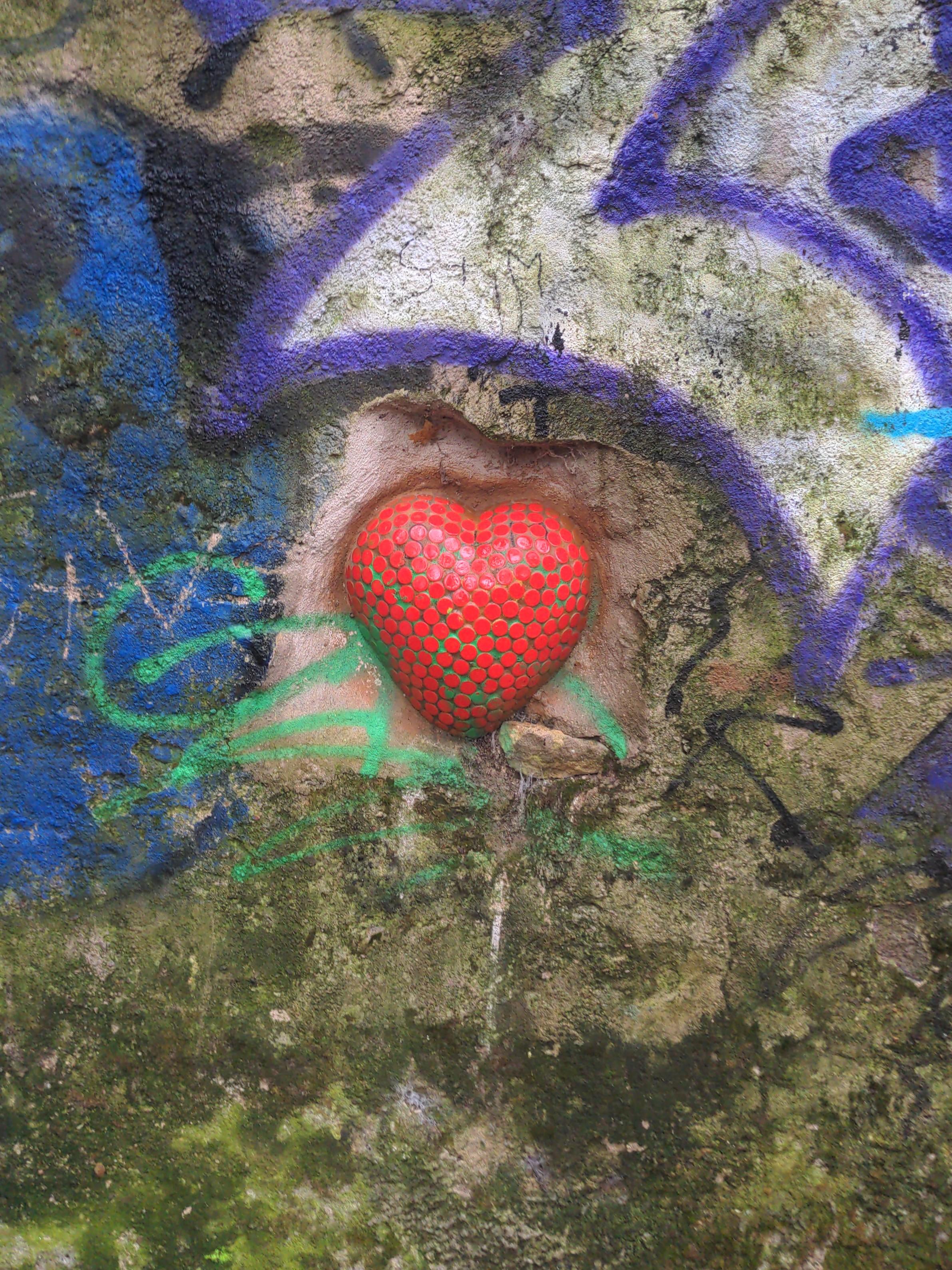 Sur un mur couvert de mousse et de graffitis, un cœur en relief fait de pastilles rouges.