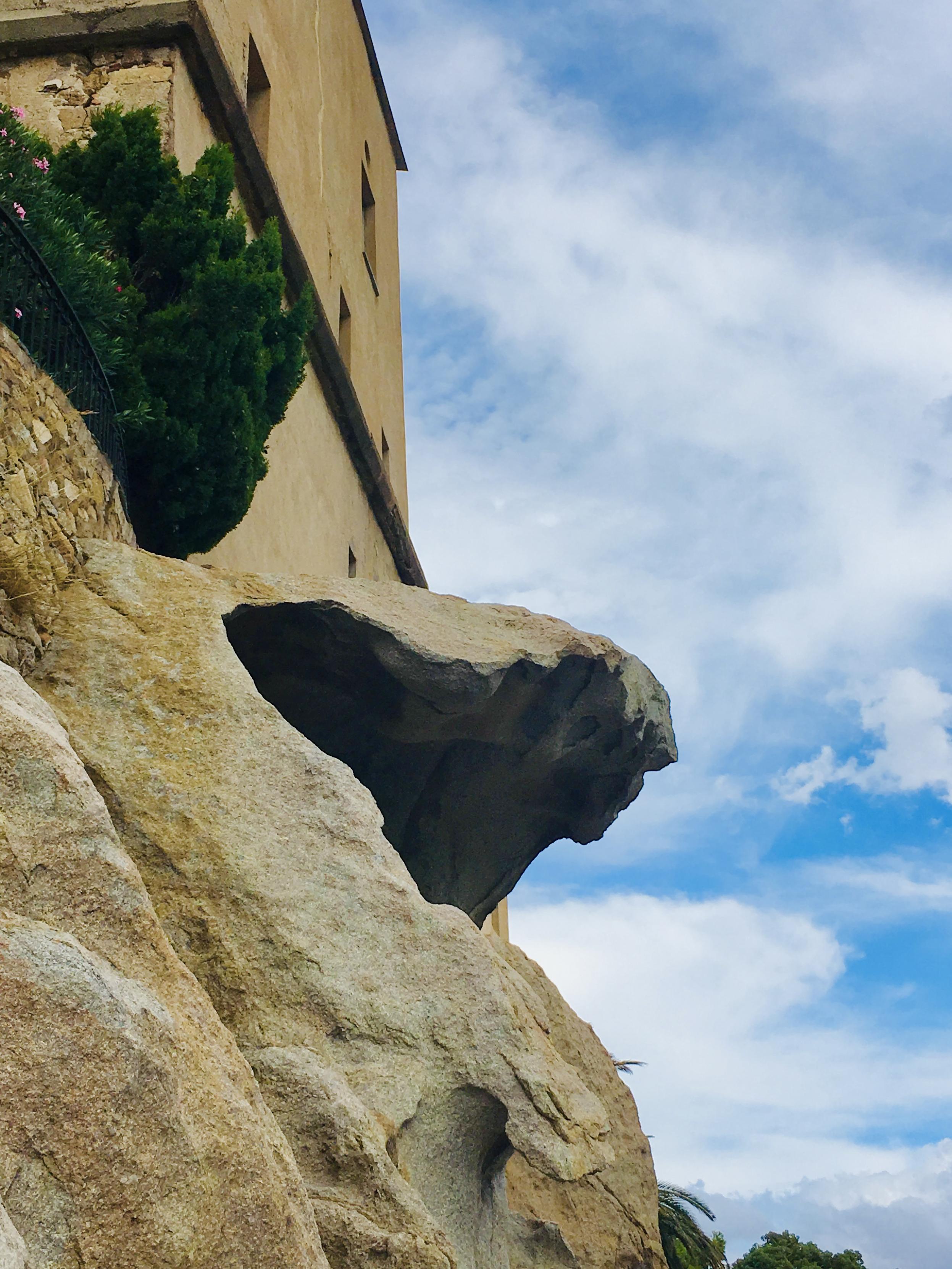 Sur la moitié gauche, une maison à quatre fenêtres est perchée sur une falaise de granit. Au-dessous de son mur d'enceinte, en haut et à gauche, se trouvent un cyprès et un laurier rose. Le rocher présente une avancée qui forme une vague de granit. La roche a des tons ocre à gris Le ciel est très bleu au milieu de nuages blancs et vaporeux.