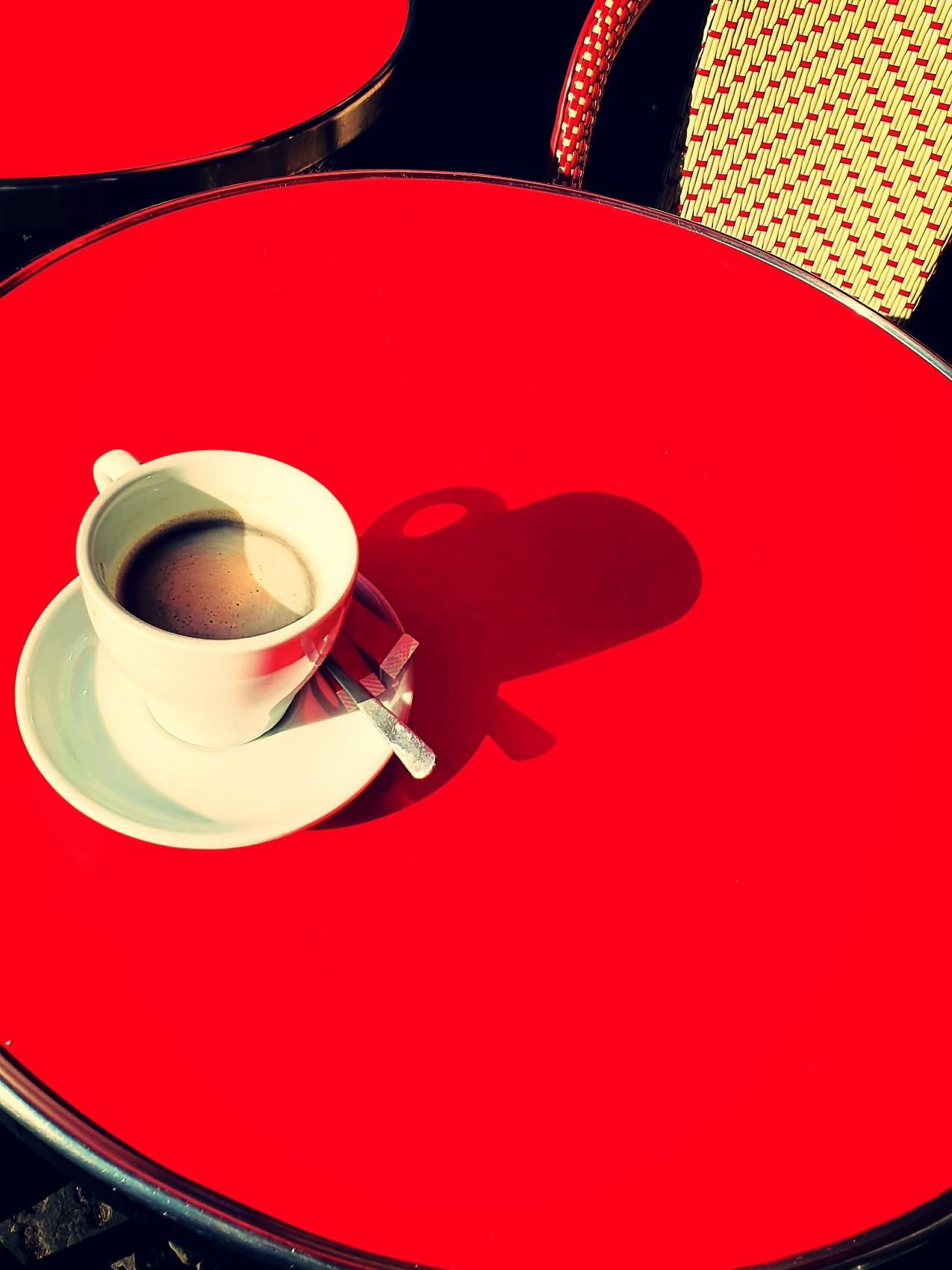Une tasse de café sur une table de bistrot parisien au soleil. Vu l'ombre de.la tasse, nous sommes.en automne/hiver, fin d'après-midi. La tasse est blanche et la table d'un rouge très éclatant. Photo de Paul depuis Bsky