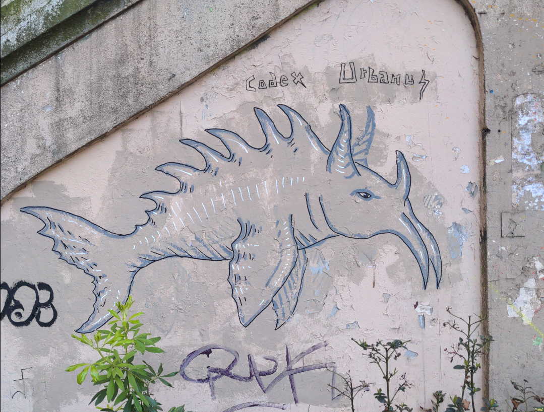 Sur un mur en béton, dans une large et peu profonde niche en biais, titré "Codex urbanus", une créature en traits bleus et noir, ayant un corps de poisson et une tête de mammifère avec deux cornes sur la tête, une sur le front et deux sur le menton.