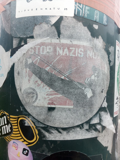 "Stop nazis now" sticker seen through what remains of a bigger sticker placed on top of it. Seen on a street pole in Helsinki.