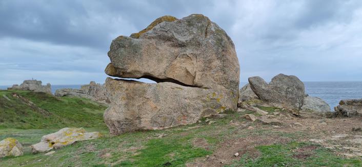 sous un ciel couvert, au pied du phare de Creac'h à Ouessant, un gros rocher granitique fendu horizontalement, évoquant une tête de dinosaure. il est couvert de lichen.