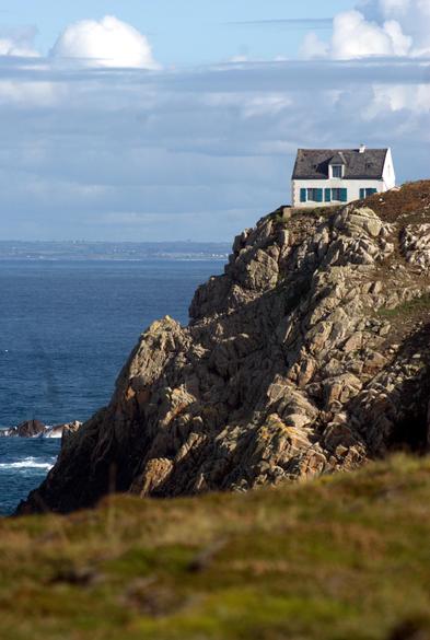 Maison blanche aux volets bleus et toit en ardoises accrochée à la falaise abrupte, sur la pointe du Stiff à Ouessant. Photo prise depuis la falaise d'en face dont on voit la végétation rase au premier plan. La falaise de la maison plonge 50 mètres plus bas dans la mer d'Iroise. La mer est peu agitée, on voit le continent à l'horizon. Temps ensoleillé avec des formations nuageuses